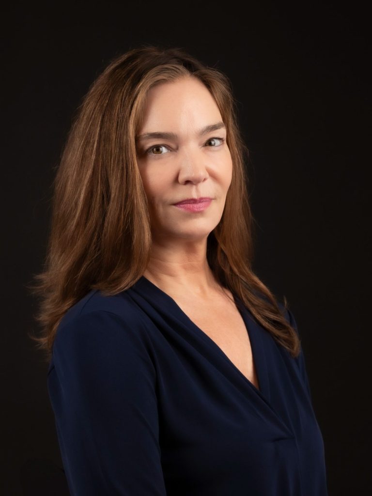 Executive woman with long, wavy hair wearing a dark blue top, smiling against a black background at Headshot Studio Portraits