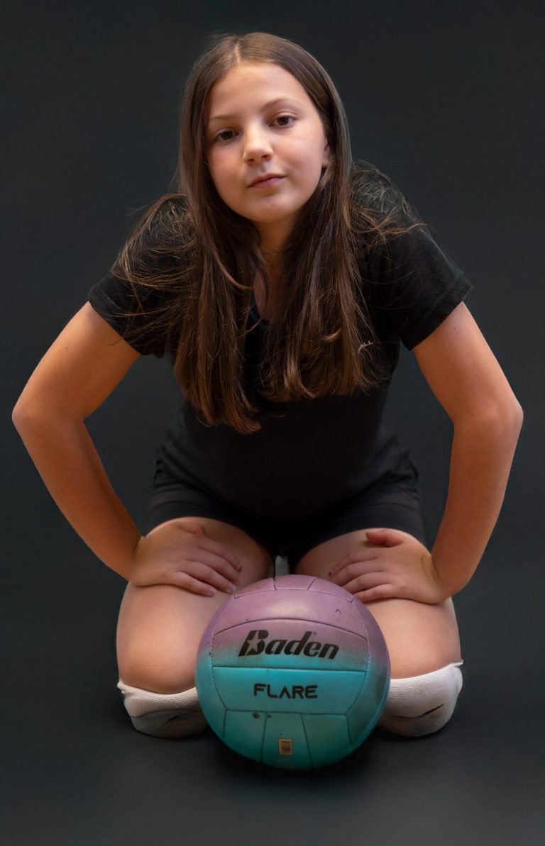 A young girl kneeling with a volleyball, looking confidently at the camera.