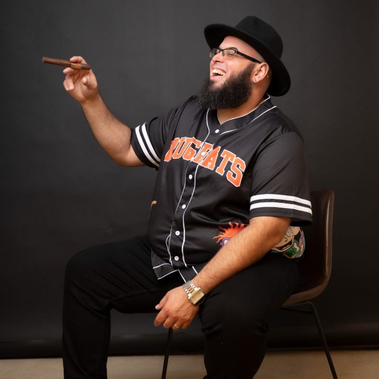 Kris Ramil in a black hat and baseball jersey siting and holding a cigar and laughing at Headshot Studio Portraits.