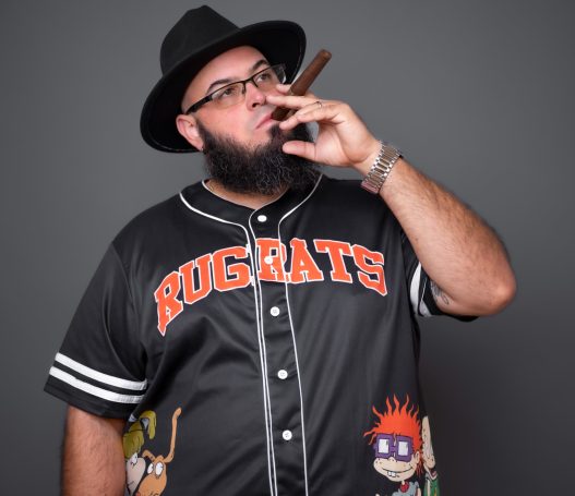 Person wearing a black jersey with cartoon characters, holding a cigar and wearing a hat.