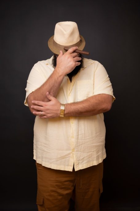 A bearded Kris Ramil at Headshot Studio Portraits in a hat poses casually, covering part of his face with a cigar .